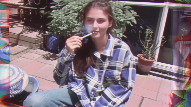 A woman wearing a plaid shirt sitting outside, holding a marijuana joint. The image has a distorted, glitched effect with bright colors on the edges. In the style of 90s camcorder footage.